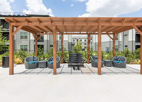 A wooden pergola with wicker chairs and a table in front of apartment buildings.