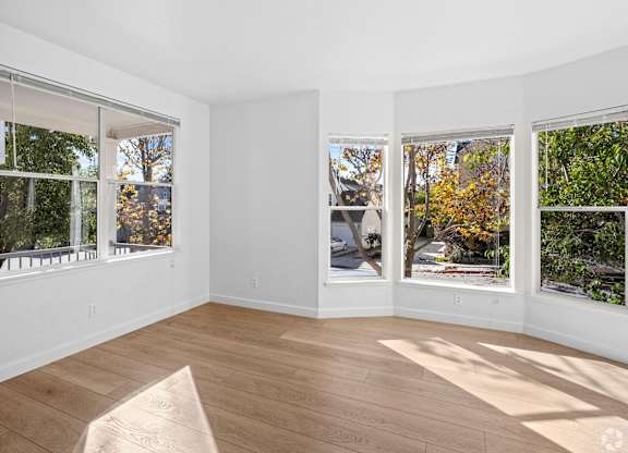 A sunny room with a wooden floor and three windows showing trees and a street at Westmont Place Townhomes Apartments, Santa Cruz, CA, 95060
