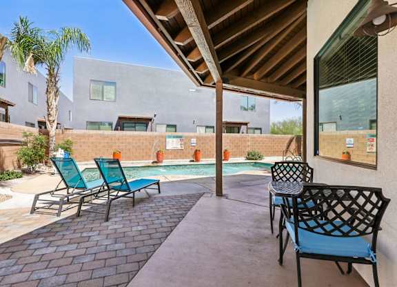 A patio with chairs and a table is surrounded by a pool and buildings.