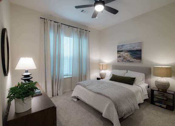 A bedroom with a bed, two lamps, a plant, and a painting on the wall at Hudson at Renaissance Apartments, Wilmington, NC 28405