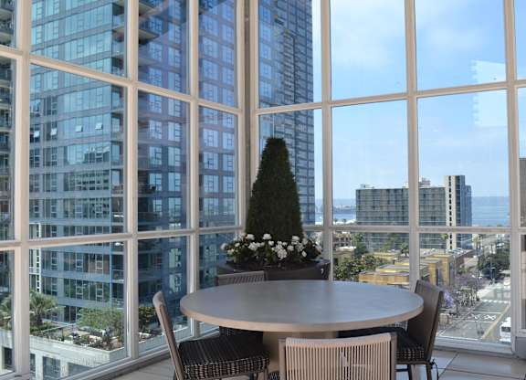 A round table with chairs and a tree in a pot is in front of a large window with a view of the city.