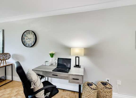 A black chair sits in front of a laptop on a desk with a clock on the wall behind it.