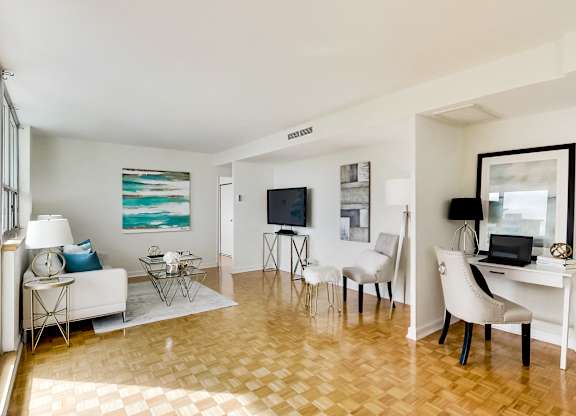 A living room with a white couch, a white chair, a black chair, a white desk, a television, a laptop, a lamp, a painting, and a floor lamp.