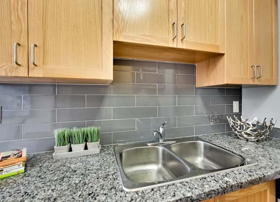 a kitchen sink with wood cabinets and gray tiles