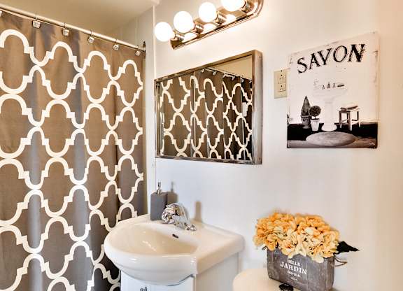 A bathroom with a patterned shower curtain, a white sink, and a framed picture of a kitchen scene.