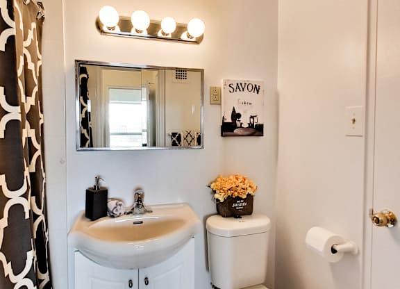 A bathroom with a white sink and toilet, a mirror with lights above it, and a sign that says