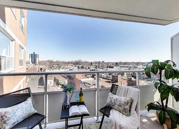 A balcony with a black chair, a black cushion, a black pillow, a white cushion, a white blanket, a green plant, a vase with flowers, a bottle, a book, and a