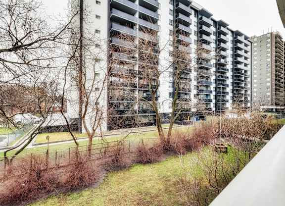 A tall residential building with balconies is surrounded by a grassy area and leafless trees.