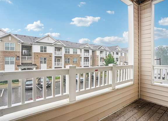 A balcony with a white railing overlooks a parking lot and apartment buildings.