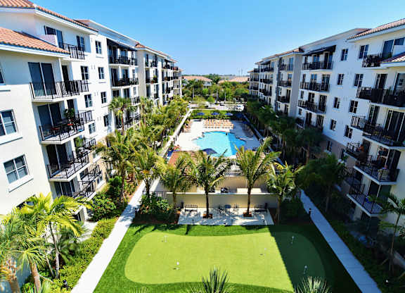 A view of an apartment complex with a pool surrounded by palm trees and a green lawn.