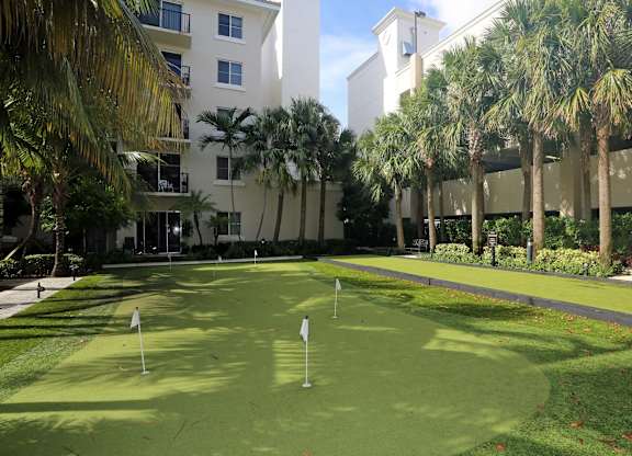 A putting green is surrounded by palm trees and a white building.