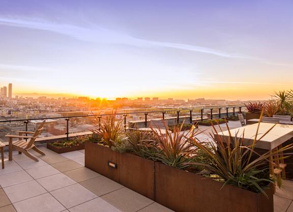 A rooftop patio with a bench and a table with a view of the city at sunset.