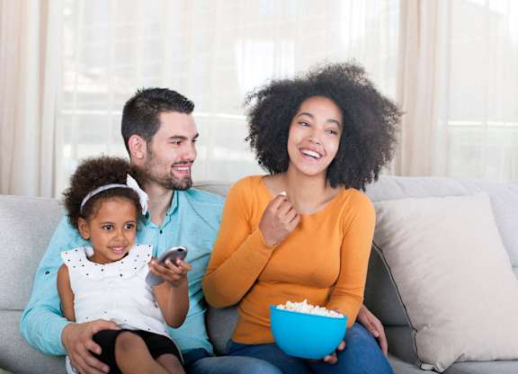 A family of three is sitting on a couch, with the woman holding a bowl of popcorn and the girl holding a remote control.