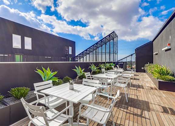 A patio with white chairs and tables is surrounded by green plants.