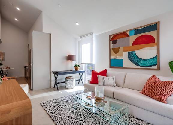 A modern living room with a white sofa, a glass coffee table, and a colorful abstract painting on the wall.