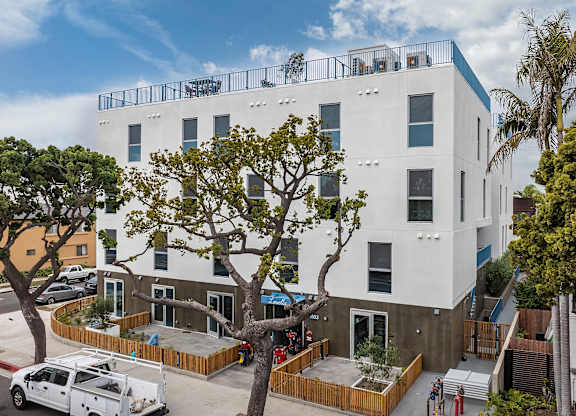 A white building with a balcony on the top floor is surrounded by trees and has a white van parked in front.