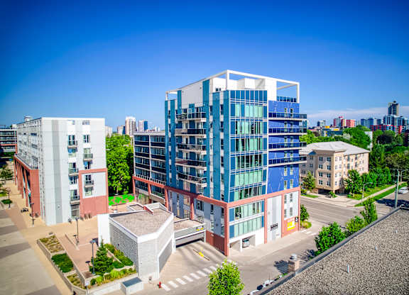 A modern multi-story building with blue and white panels is surrounded by other buildings and greenery.