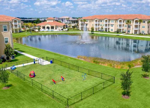 An apartment community surrounding a pond with a fountain in the center next to a dog park that's a fenced-in grassy area.