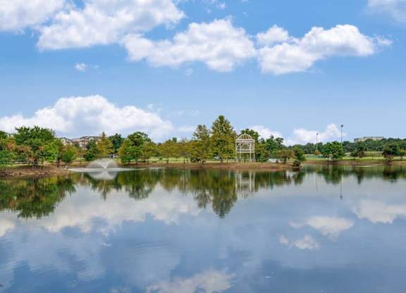 A serene lake with a fountain in the middle, surrounded by trees and a clear blue sky.