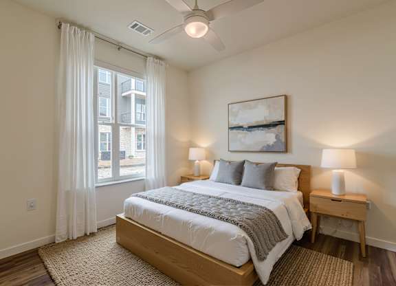 A bedroom with a large bed, a ceiling fan, and a window with white curtains.