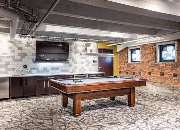 A pool table is in the middle of a room with a brick wall and a television.