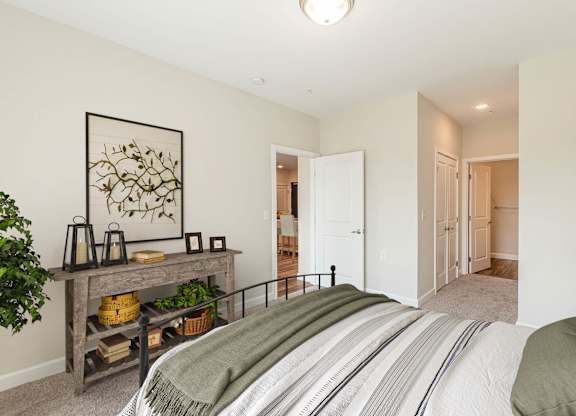A bedroom with a bed, a table with a plant, and a painting on the wall. at Avery Landing Apartments, Greenville, 29605