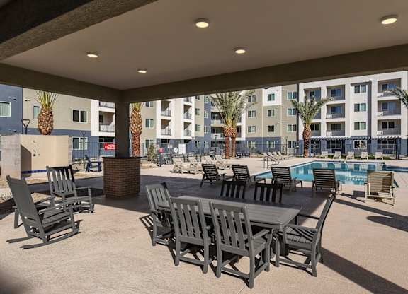 A patio with chairs and a table is in front of a pool and apartment building.