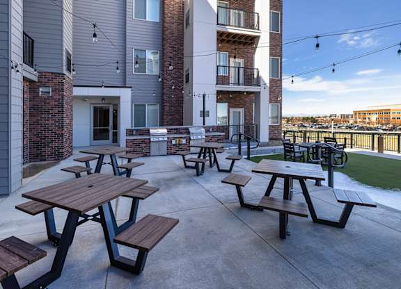 Picnic tables and benches are set up on a patio in front of apartment buildings.