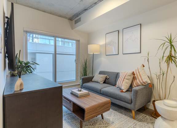 A living room with a grey couch, a wooden coffee table, a white toilet, and a large window.