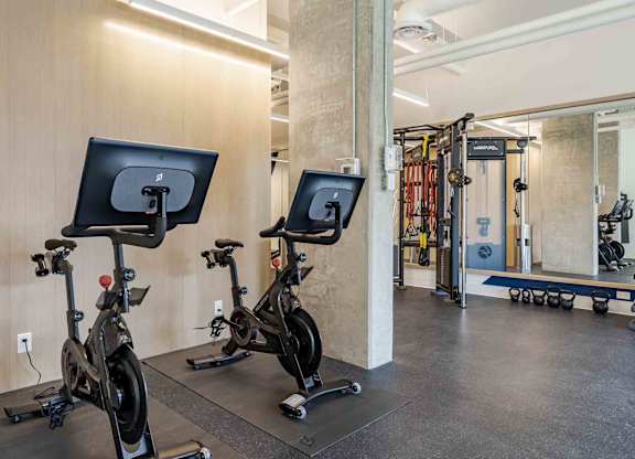Two stationary bikes are in a gym with a mirror and weights in the background.