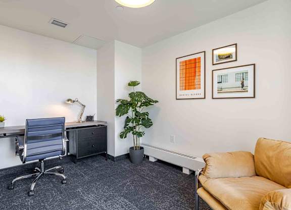 A room with a grey chair, a desk, a potted plant, and two framed pictures on the wall.