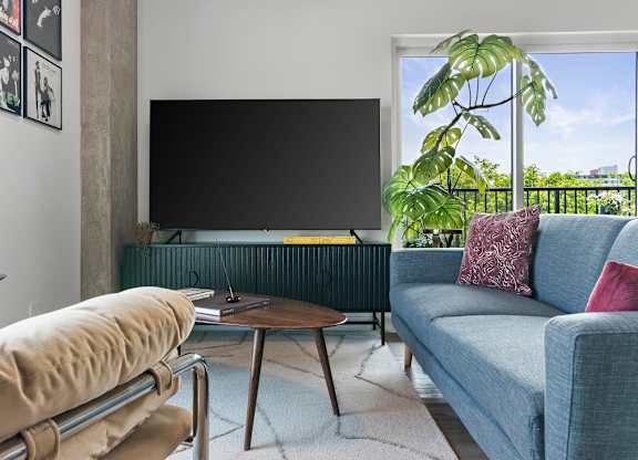 A living room with a grey couch, a brown chair, a black television and a window with a view of the city.