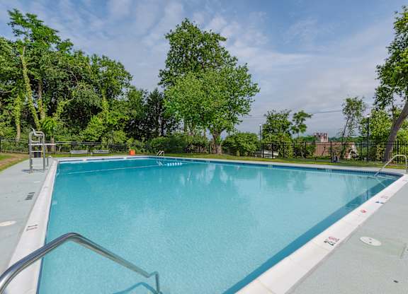 A rectangular blue swimming pool surrounded by a metal fence and trees.