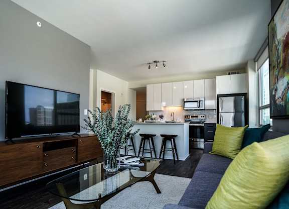 A modern living room with a television, a glass coffee table, and a kitchen in the background.