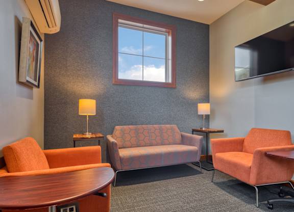 A living room with a grey carpet and a grey wall with a window and a flat screen TV.