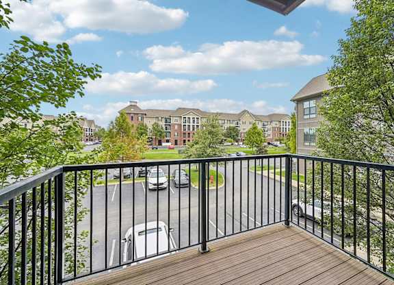 A balcony with a black railing overlooks a parking lot and buildings.