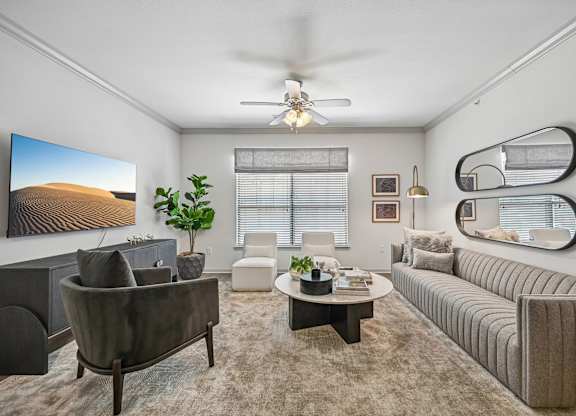A living room with a grey sofa, a grey armchair, a coffee table, a potted plant, a painting, a mirror, and a ceiling fan.