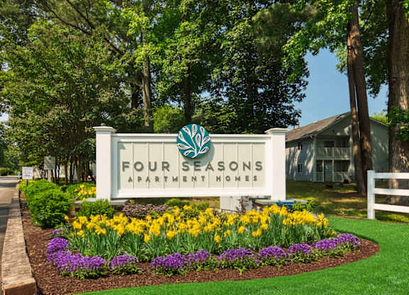 A sign for Four Seasons Apartment Homes is surrounded by flowers and greenery.