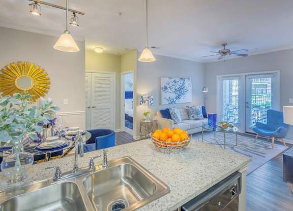 A kitchen with a sink, a counter, a fridge, a fan, a chair, a table, a vase, a bowl, and a window.