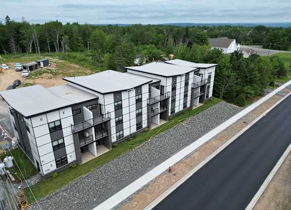 A modern building with a grey and white exterior is surrounded by a gravel area and a paved road.