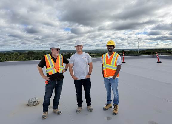 Three construction workers wearing safety vests and hard hats stand on a rooftop.