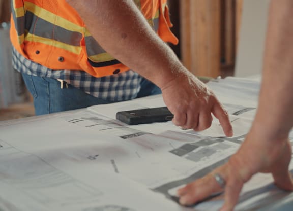 A person in an orange vest is pointing at a blueprint.