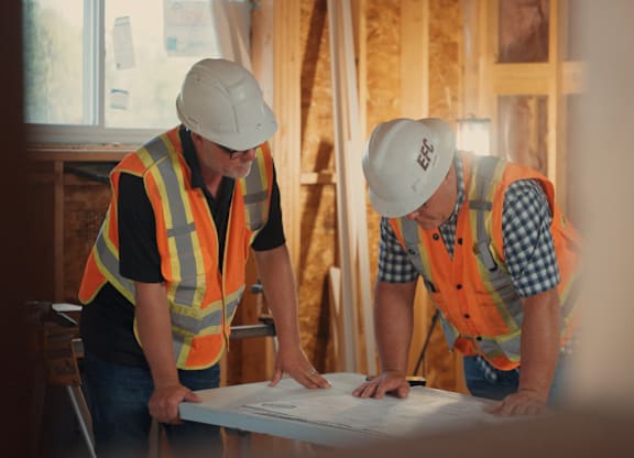 Two construction workers wearing safety helmets and vests are examining a blueprint on a table.