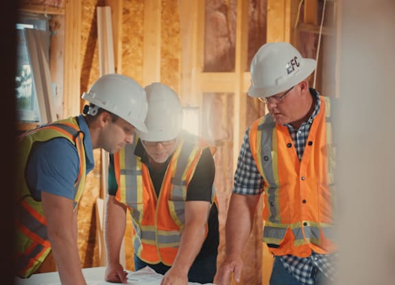 Three construction workers wearing safety helmets and vests are looking at a blueprint.