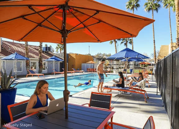 A woman is working on her laptop under an orange umbrella at a poolside table.