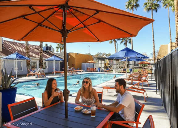 Three people are sitting at a table under an orange umbrella by a pool.