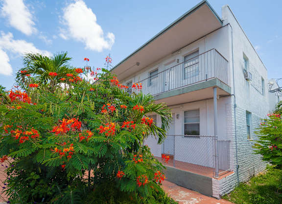 A white house with a balcony and a tree with red flowers in front.