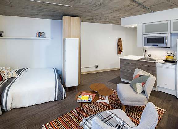 A bedroom with a bed, chair, and a kitchen area.