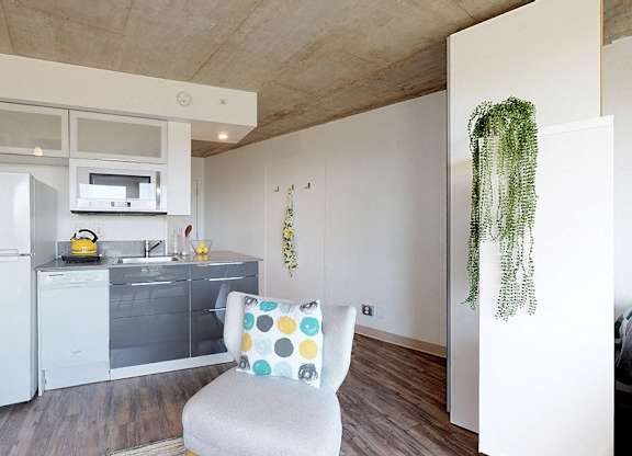 A white chair with a multicolored pillow sits in a kitchen with a white refrigerator and white cabinets.
