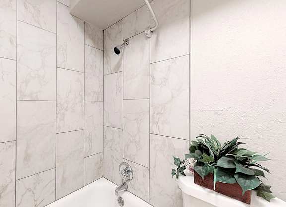 A white bathroom with a marble tile wall and a plant on the tub.